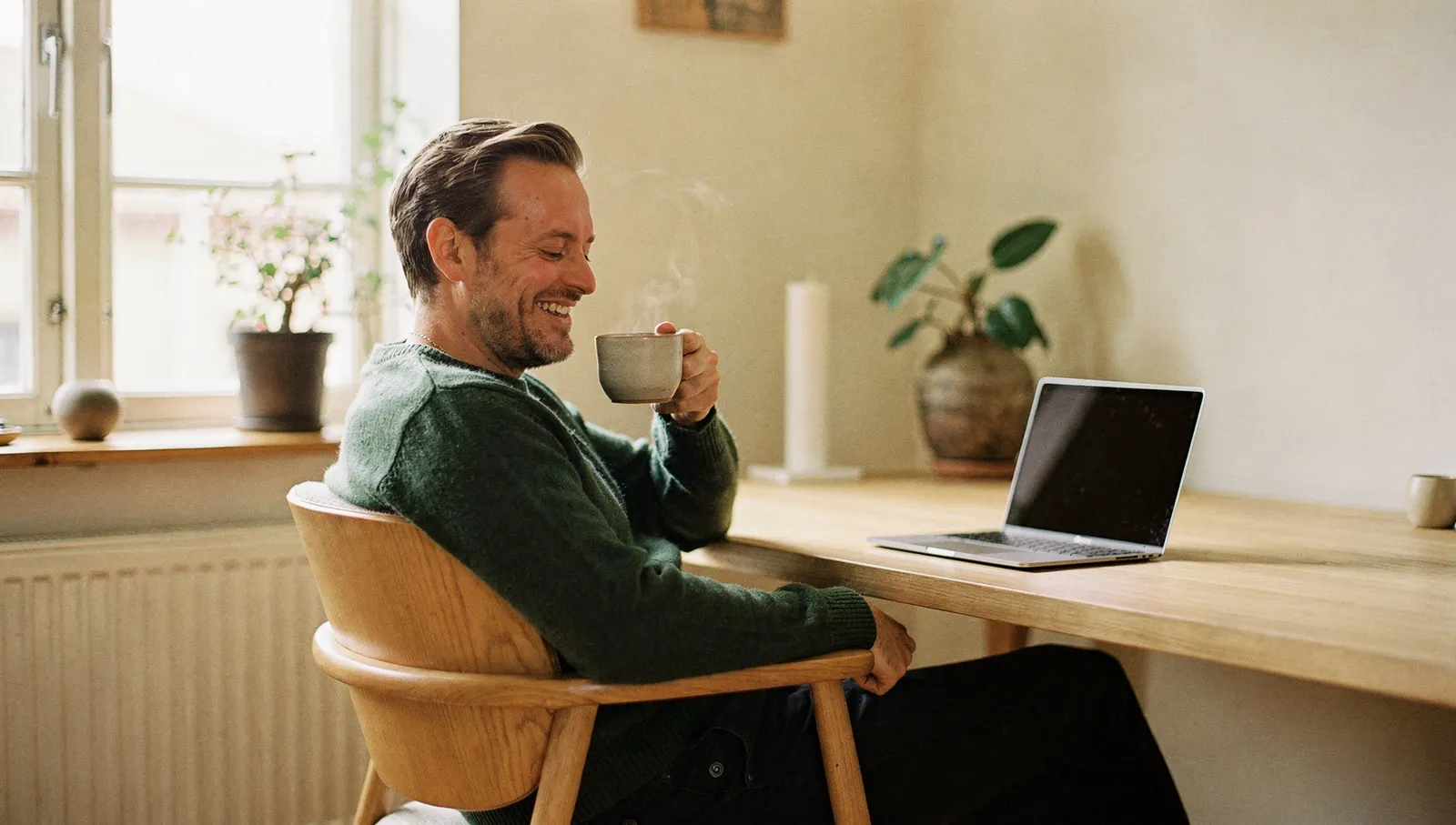 Avslappet mann lener seg tilbake med kaffekopp og smiler til laptopen på hjemmekontoret